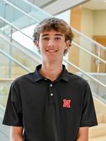 Young man in a black polo with a red "N" logo, standing in front of a staircase indoors.