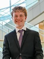 Young man in suit and tie smiling in a modern building with glass stair railings.