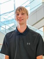 Young man wearing a black polo shirt, standing in a bright indoor space with stairs in the background.