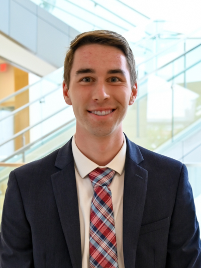 Smiling man in a suit and plaid tie, standing in front of glass stairs indoors.