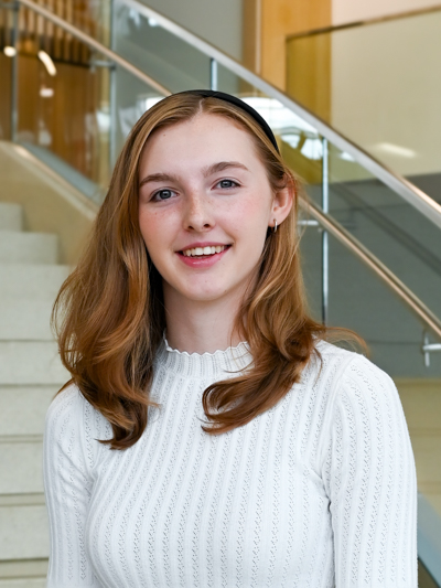 Smiling woman with red hair in a white sweater standing near stairs indoors.