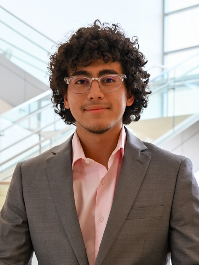 Man with curly hair and glasses, wearing a gray suit and pink shirt, standing by modern stairs.