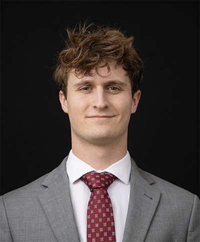 Man in a gray suit with a red tie, smiling slightly against a black background.
