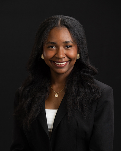Smiling woman in formal black blazer, white top, gold necklace; studio portrait on black background.