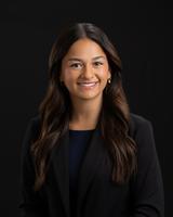 Smiling woman with long dark hair wearing a black blazer, set against a black background.