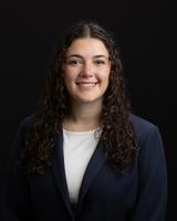 Smiling woman with curly dark hair, wearing a navy blazer and white top, against a black background.