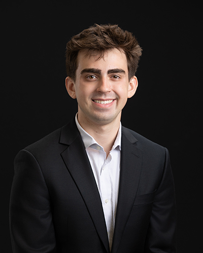 Man in a suit and collared shirt smiling against a black background.