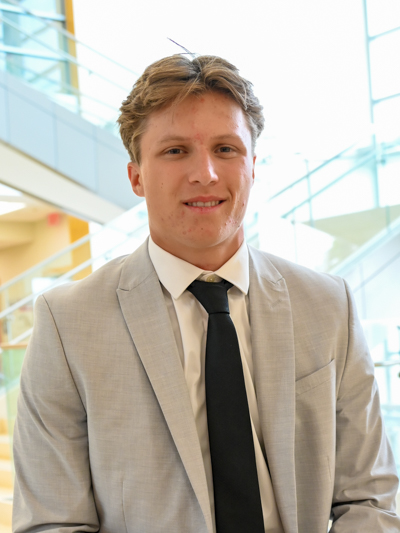 Man in light gray suit with black tie, standing in bright indoor setting with glass staircase.