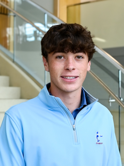 Young man in a light blue sweatshirt standing in front of a staircase indoors.