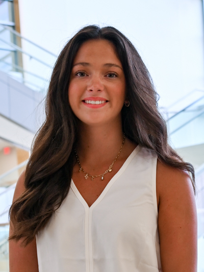 Smiling woman with long brown hair in a white top, standing indoors near glass railings.