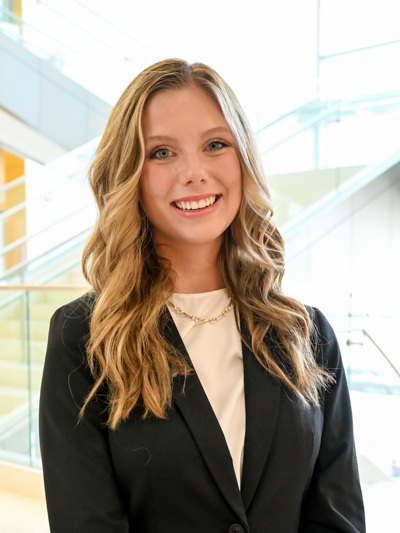 Smiling woman with wavy blonde hair in a black blazer, standing in a bright modern building.
