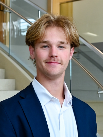 Young man with blonde hair and a mustache wearing a navy suit and white shirt, standing by stairs indoors.