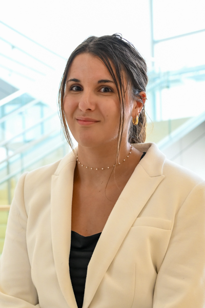 Woman wearing a white blazer, smiling softly in bright indoor space with stairs behind.