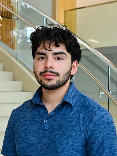 Man with curly hair and beard wearing a blue shirt, standing by stairs with glass railings indoors.