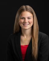Smiling woman with long brown hair wearing a black blazer and red shirt against a black background.