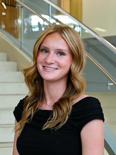 Smiling woman with wavy blonde hair in a black top, standing indoors near stairs and glass railing.