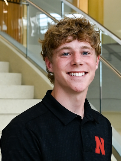 Smiling young man in a black shirt with a red letter "N," standing near a staircase indoors.