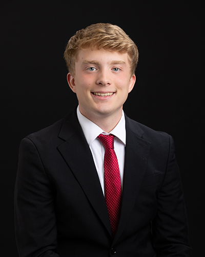 Young man in a black suit, white shirt, and red tie, smiling against a dark background.