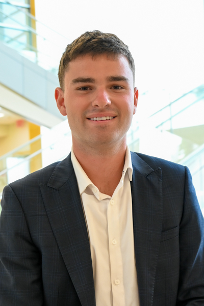 Smiling man in a dark blazer with a white shirt, sitting indoors near glass railings.