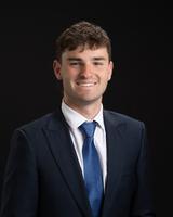Smiling man in a dark suit and blue tie against a black background.