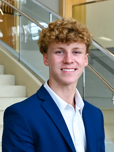 Smiling young man in a blue blazer and white shirt, standing near a staircase indoors.