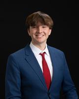 Smiling person wearing a blue suit, white shirt, and red tie against a black background.