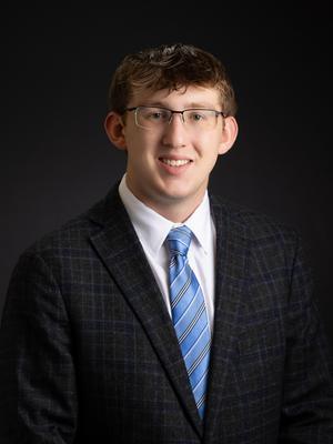 Man wearing a suit, tie, and glasses against a dark background, smiling softly.