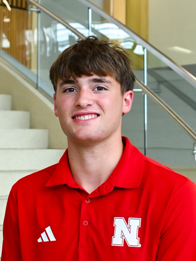 Smiling man in a red shirt with white "N" logo, indoors near stairs and glass railing.