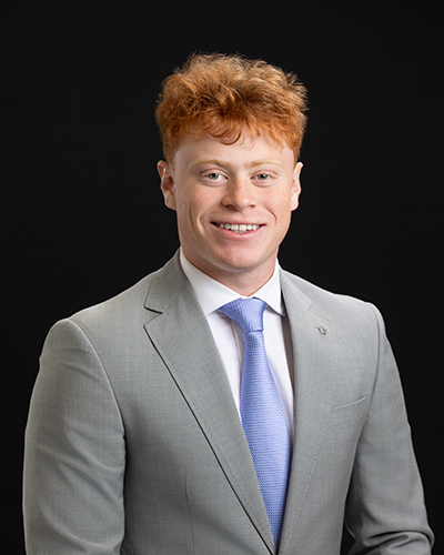 Smiling man in a gray suit and light blue tie against a black background.