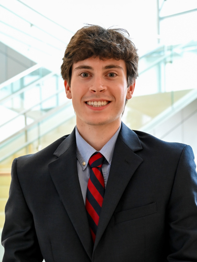 Man in a dark suit and red striped tie smiling in front of glass staircase indoors.