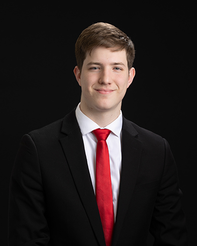 Smiling man in a black suit, white shirt, and red tie, posed against a black background.