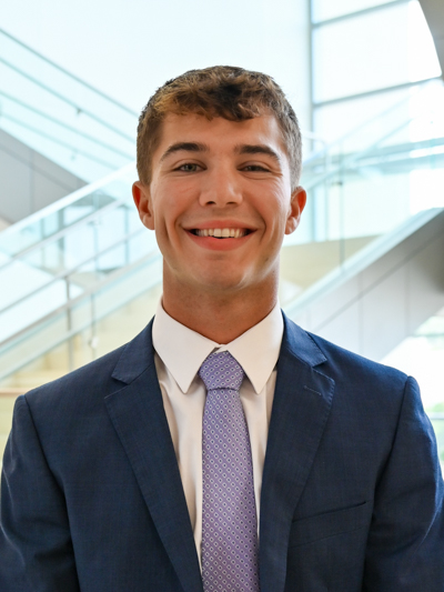 Professional man in a navy suit and lavender tie smiling in a modern office setting.