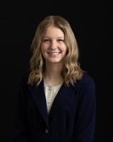 Smiling woman with wavy blonde hair in a navy blazer against a black background.