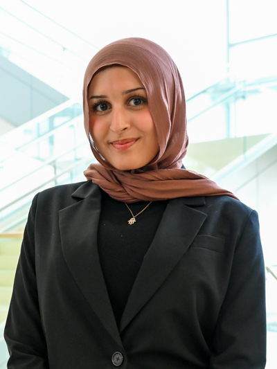 Smiling woman in a brown hijab and black blazer, standing indoors near a staircase.
