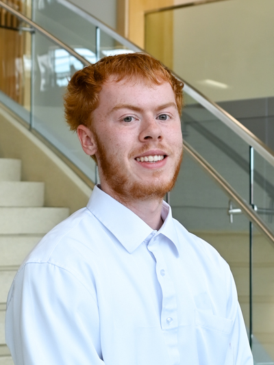 Smiling man with red hair in a white shirt, standing near a staircase with glass railing indoors.