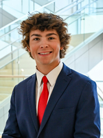 Smiling young man in a blue suit and red tie, standing in a modern glass stairwell.