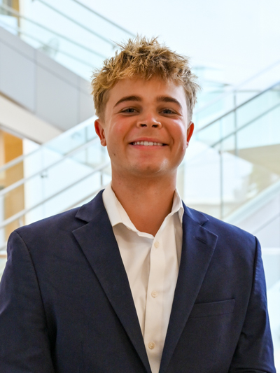 Smiling person in a navy blazer and white shirt, standing near modern glass staircases indoors.