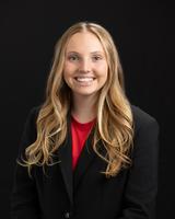 Smiling woman with long blonde hair wearing a black blazer and red top, against a black background.