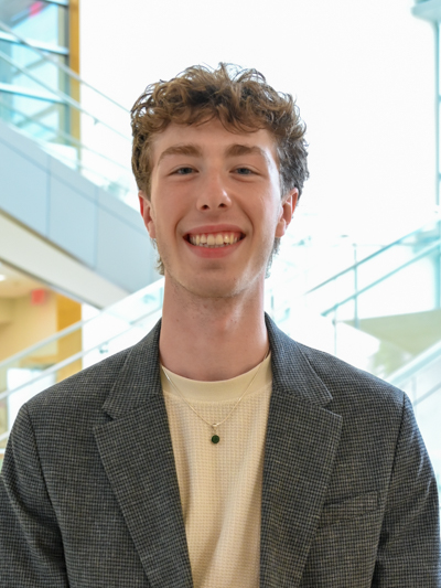 Smiling young man wearing a blazer and cream shirt, standing indoors near glass stair railing.