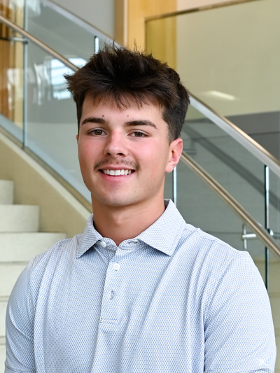 Smiling young man in a light blue shirt standing in front of stairs and glass railing indoors.