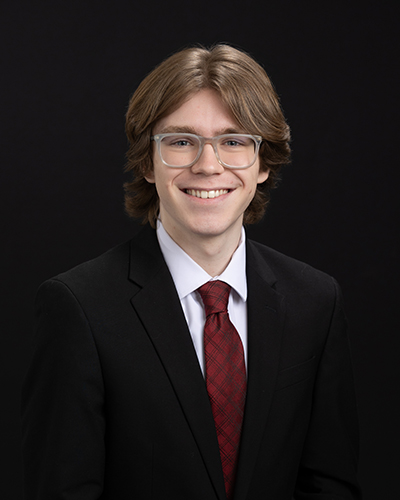 Smiling man in a suit, red tie, and glasses against a black background.