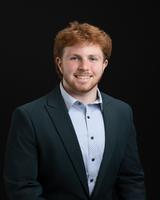 Man with red hair wearing a dark suit and light shirt, smiling against a black background.