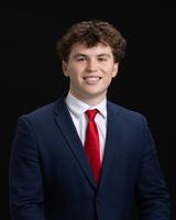 Young man in a blue suit, white shirt, and red tie, smiling against a black background.