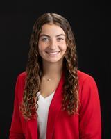 Smiling woman with long curly hair in a red blazer and white shirt, posed against a black background.