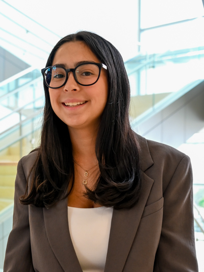 Woman with glasses smiling in a modern, brightly lit building with stairs in the background.
