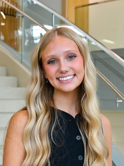 Smiling woman with long blonde hair stands near a staircase in a modern building.