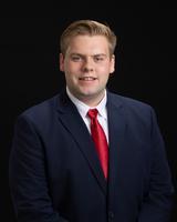 Man in a navy suit with a red tie, smiling against a black background.