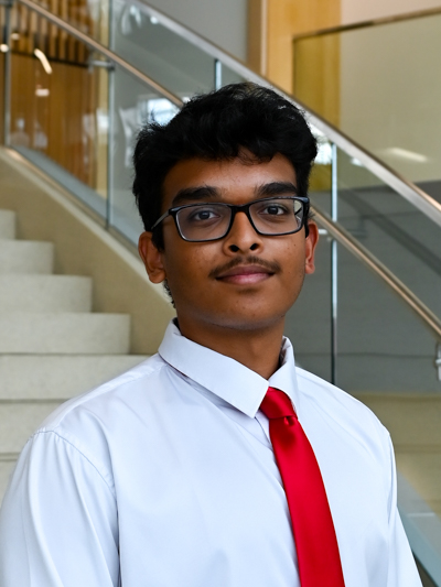 Young man in glasses wearing a white shirt and red tie, standing near stairs indoors.