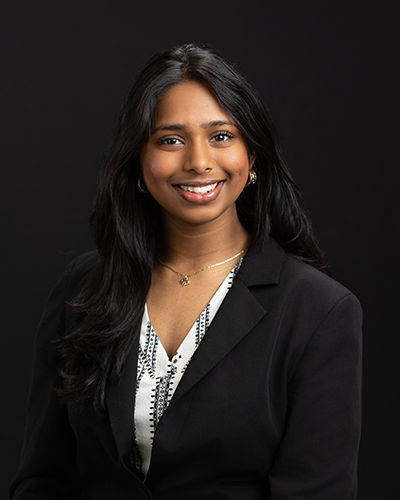 Smiling woman in a black blazer and patterned blouse against a black background.