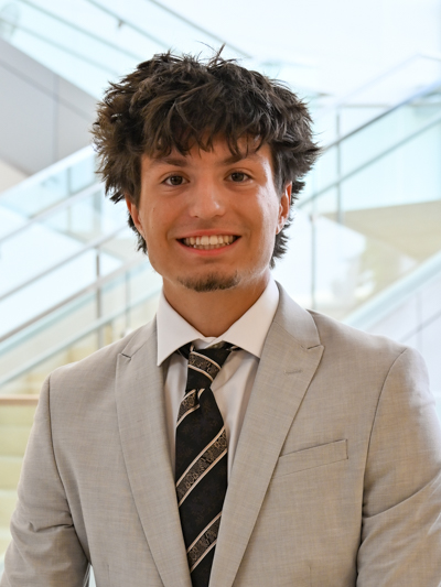 Man in a light gray suit and patterned tie, smiling in front of a modern staircase.